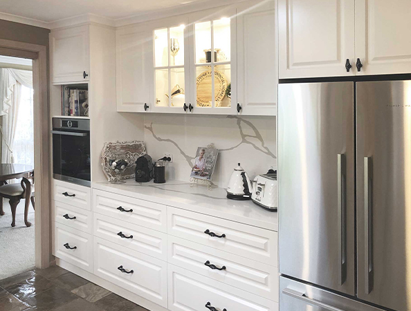 Sleek white kitchen with modern cabinets, glass display, built-in oven, and stainless steel fridge. Dining room visible.