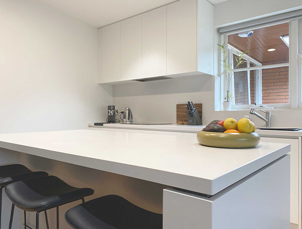 Modern kitchen with white cabinetry, a sleek island, stools, and a bowl of fruit on the counter.