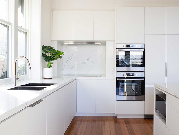 Minimalist white kitchen with wood floor, built-in ovens, and a potted plant by a window. Minimalist white kitchen with wood floor, built-in ovens, and a potted plant by a window.