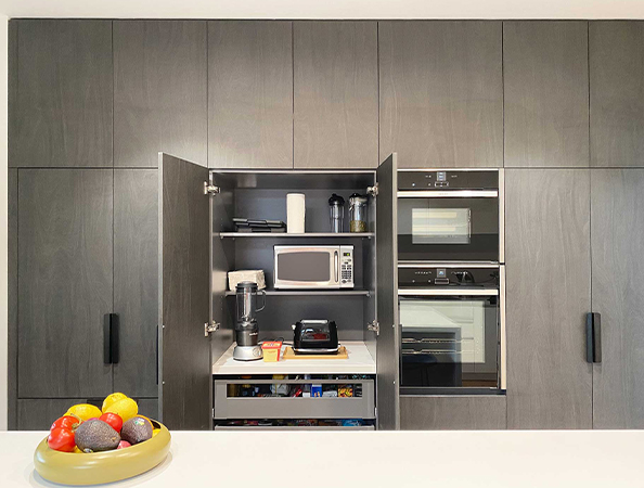 Modern kitchen with open cabinets showing a microwave, blender, and stacked ovens; fruit bowl on the countertop.