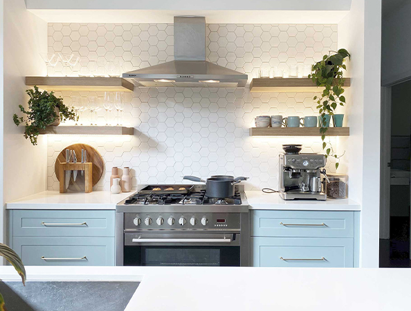 Modern kitchen with a hexagonal tile backsplash, blue cabinets, a stove, shelves with plants, dishes, and a coffee maker.