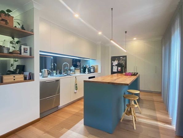 Modern kitchen with a wooden island, blue base, bar stools, white cabinets, and plants on shelves. Natural light from windows.
