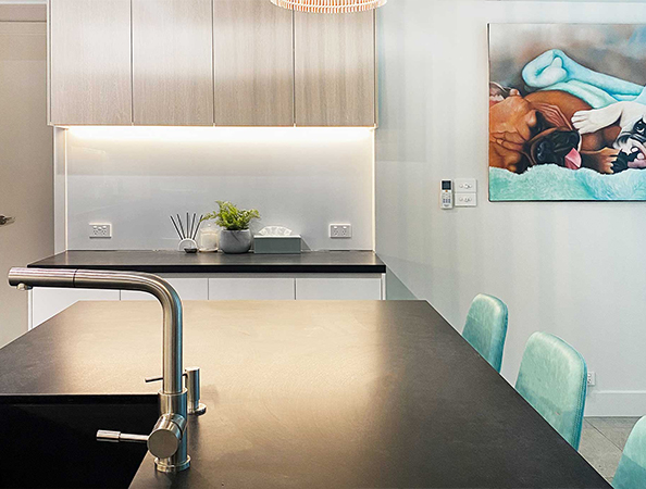 Modern kitchen with black countertop, stainless steel faucet, light wood cabinets, and a colorful wall painting.
