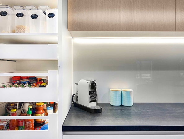 Modern kitchen with a coffee machine, blue mugs, and an open pantry showing jars and canned goods.