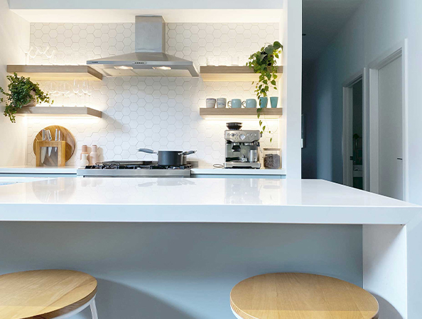 Modern kitchen with white countertops, hexagonal tile backsplash, wooden shelves, plants, and light wood stools.