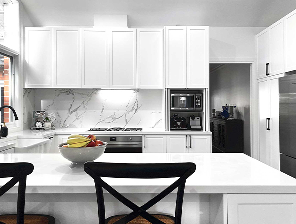 Modern white kitchen with marble backsplash, fruit bowl on counter, and black chairs. Appliances include oven and microwave.