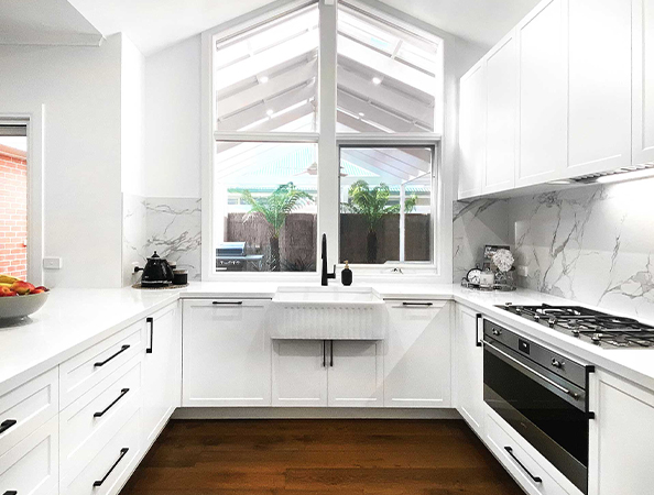 Bright kitchen with white cabinets, marble backsplash, farmhouse sink, and large window. Wood floor and skylight roof.