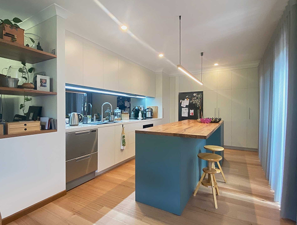 Modern kitchen with a teal island, wooden stools, and plants on shelves. Sunlight streams through sheer curtains.