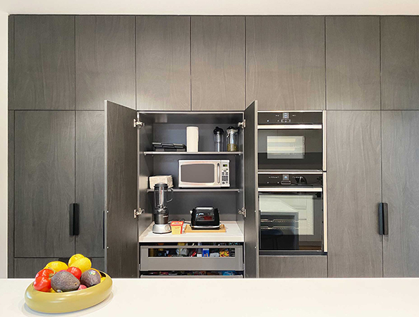 Modern kitchen cabinet with open doors revealing appliances. Bowl of fruit on the counter in the foreground.