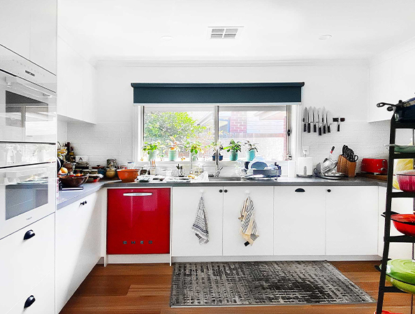 Bright kitchen with white cabinets, red dishwasher, large window, knives on wall, and a rug on wooden floor.