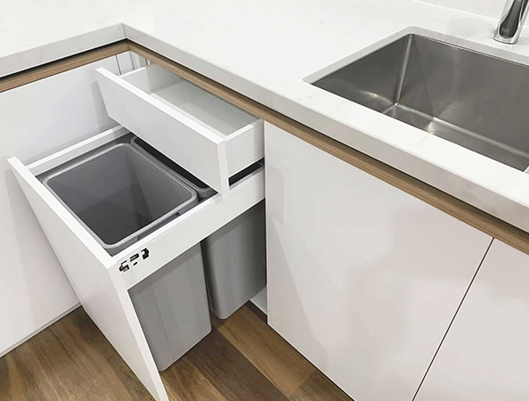 Modern kitchen with a pull-out trash and recycling bin drawer, adjacent to a stainless steel sink and wooden floor.