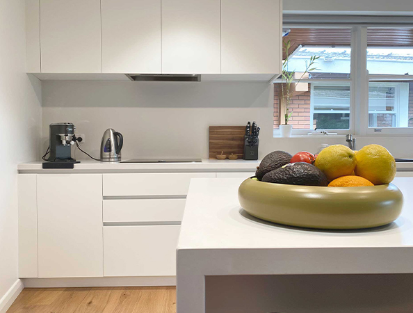Modern kitchen with white cabinets, a countertop appliance, and a bowl of fruits on the island. Natural light from window.