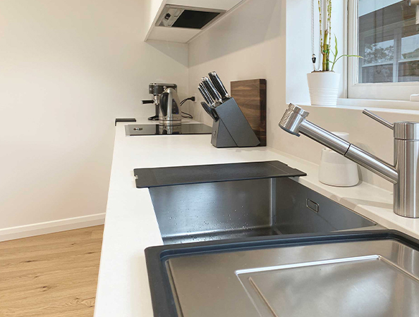 Modern kitchen with white countertops, stainless steel sink, knife block, coffee maker, and a small potted plant.