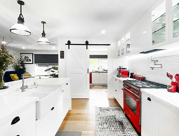 Modern kitchen with red stove, white cabinets, farmhouse sink, and sliding barn door, illuminated by pendant lights.