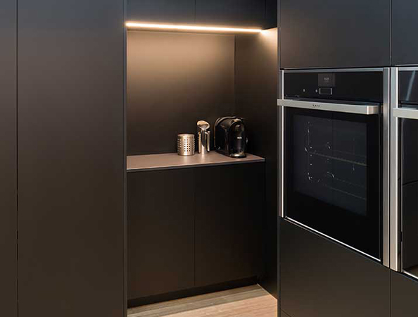 Modern kitchen with black cabinets, built-in oven, under-cabinet lighting, and a small coffee station.