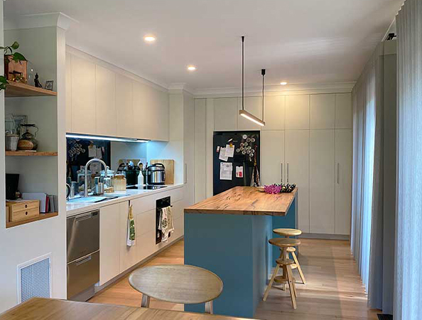 Modern kitchen with a wooden island, stools, and appliances. Refrigerator with magnets. Warm lighting and wooden floor.