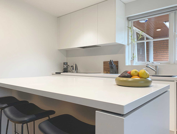 Modern kitchen with white countertops, black stools, fruit bowl, and window.