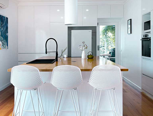 Modern kitchen with a white island, black faucet, and three white stools. Wooden floor and large window in background.