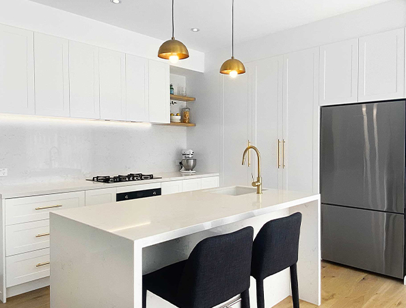 Modern kitchen with white cabinets, island with two black chairs, gold fixtures, and stainless steel fridge.