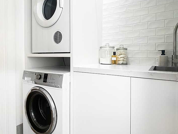 Modern laundry room with stacked washer and dryer beside a white counter and sink, jars on counter.
