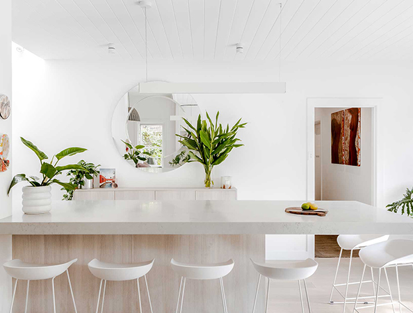 Minimalist white kitchen with a large mirror, green plants, and modern white barstools around a marble island. Minimalist white kitchen with a large mirror, green plants, and modern white barstools around a marble island.