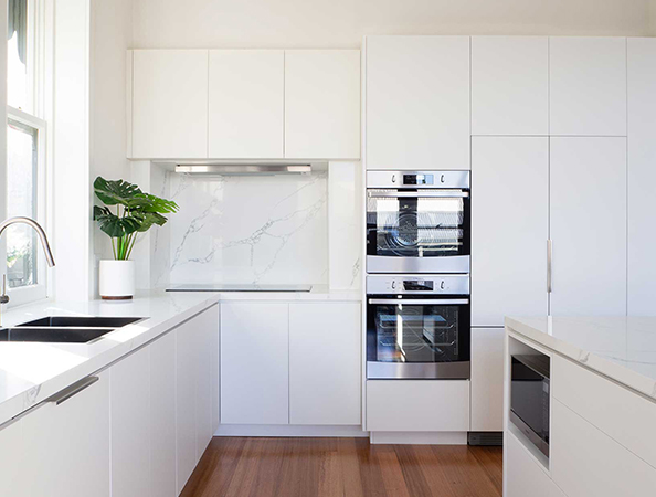 Modern white kitchen with built-in ovens, a potted plant, and wooden flooring. Bright and minimalist design.