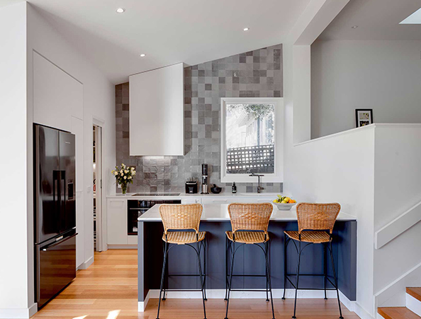 Modern kitchen with a black fridge, gray tiled backsplash, and island with wicker stools. Wood flooring and white walls.