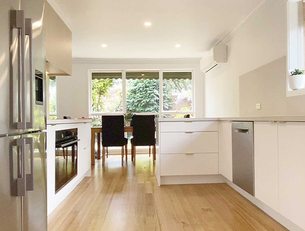 Modern kitchen with white cabinets, wooden floor, stainless steel appliances, and a dining table by large windows.