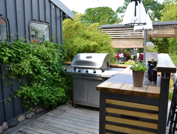 Outdoor patio with a stainless steel grill, wooden deck, lush greenery, and a wooden bar with potted plants. Outdoor patio with a stainless steel grill, wooden deck, lush greenery, and a wooden bar with potted plants.