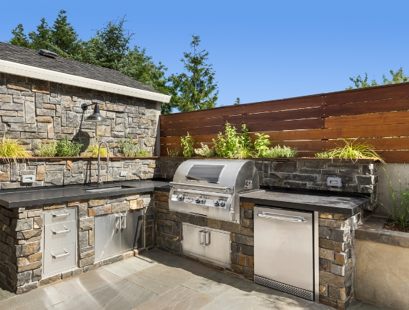 Outdoor kitchen with stone counters, grill, plants, and wooden fence on a sunny day. Outdoor kitchen with stone counters, grill, plants, and wooden fence on a sunny day.