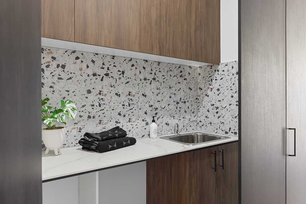 Modern laundry room with terrazzo backsplash, wooden cabinets, a small sink, black towels, and a potted plant. Modern laundry room with terrazzo backsplash, wooden cabinets, a small sink, black towels, and a potted plant.