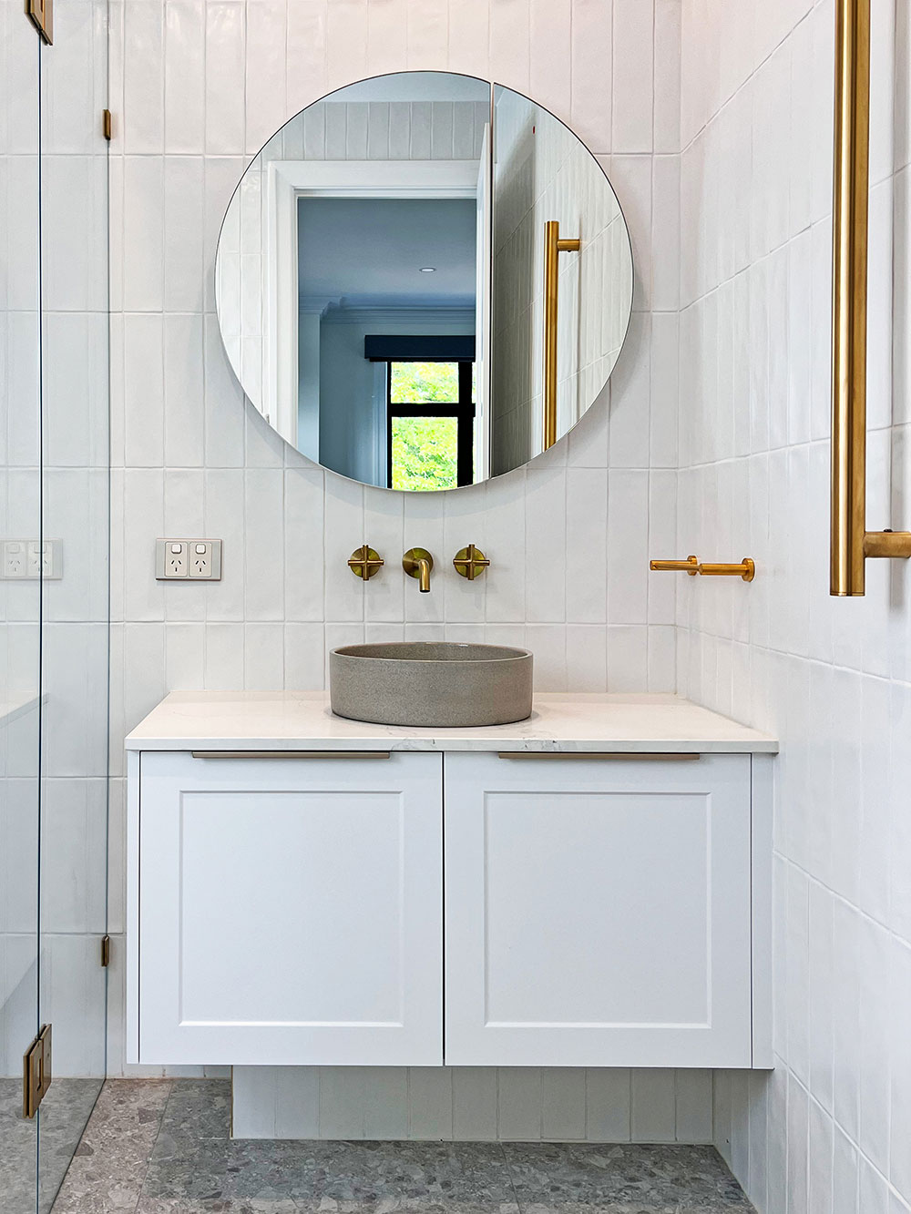 Modern bathroom with round mirror, white vanity, gray basin, and gold fixtures. Natural light from window. Modern bathroom with round mirror, white vanity, gray basin, and gold fixtures. Natural light from window.