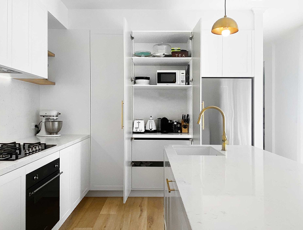 Modern white kitchen with open cupboard showing appliances, island sink with gold faucet, and pendant light overhead. Modern white kitchen with open cupboard showing appliances, island sink with gold faucet, and pendant light overhead.