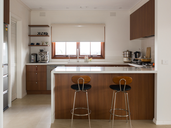 Modern kitchen with wooden cabinets, two stools at the counter, and appliances near the window with a roller blind.