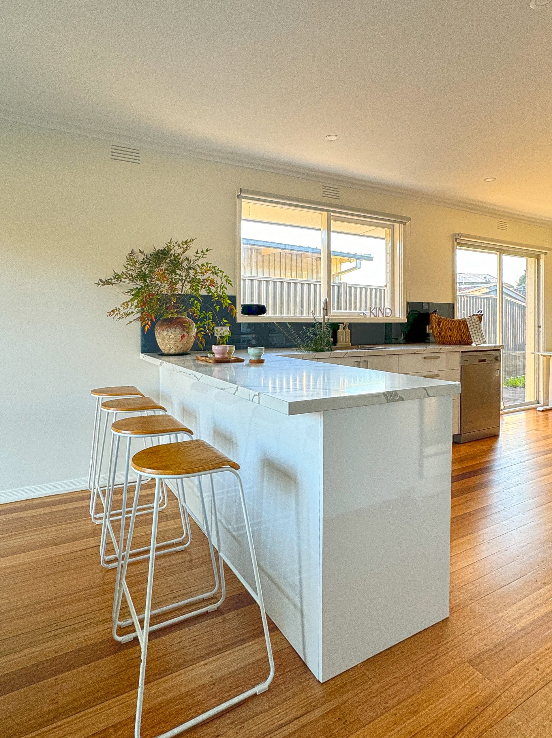 Modern kitchen with a white island, four wooden stools, potted plant, and large windows letting in natural light. Modern kitchen with a white island, four wooden stools, potted plant, and large windows letting in natural light.