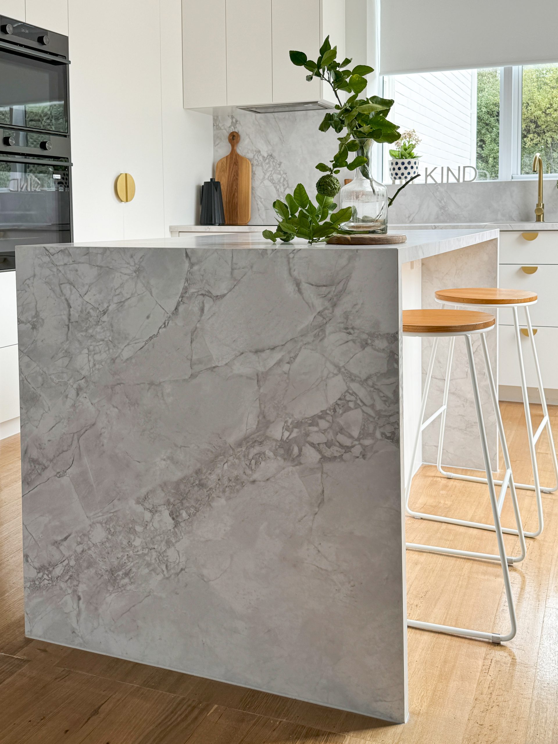 Modern kitchen with a marble island, wooden stools, plant, and decorative sign in a bright, minimalist setting. Modern kitchen with a marble island, wooden stools, plant, and decorative sign in a bright, minimalist setting.