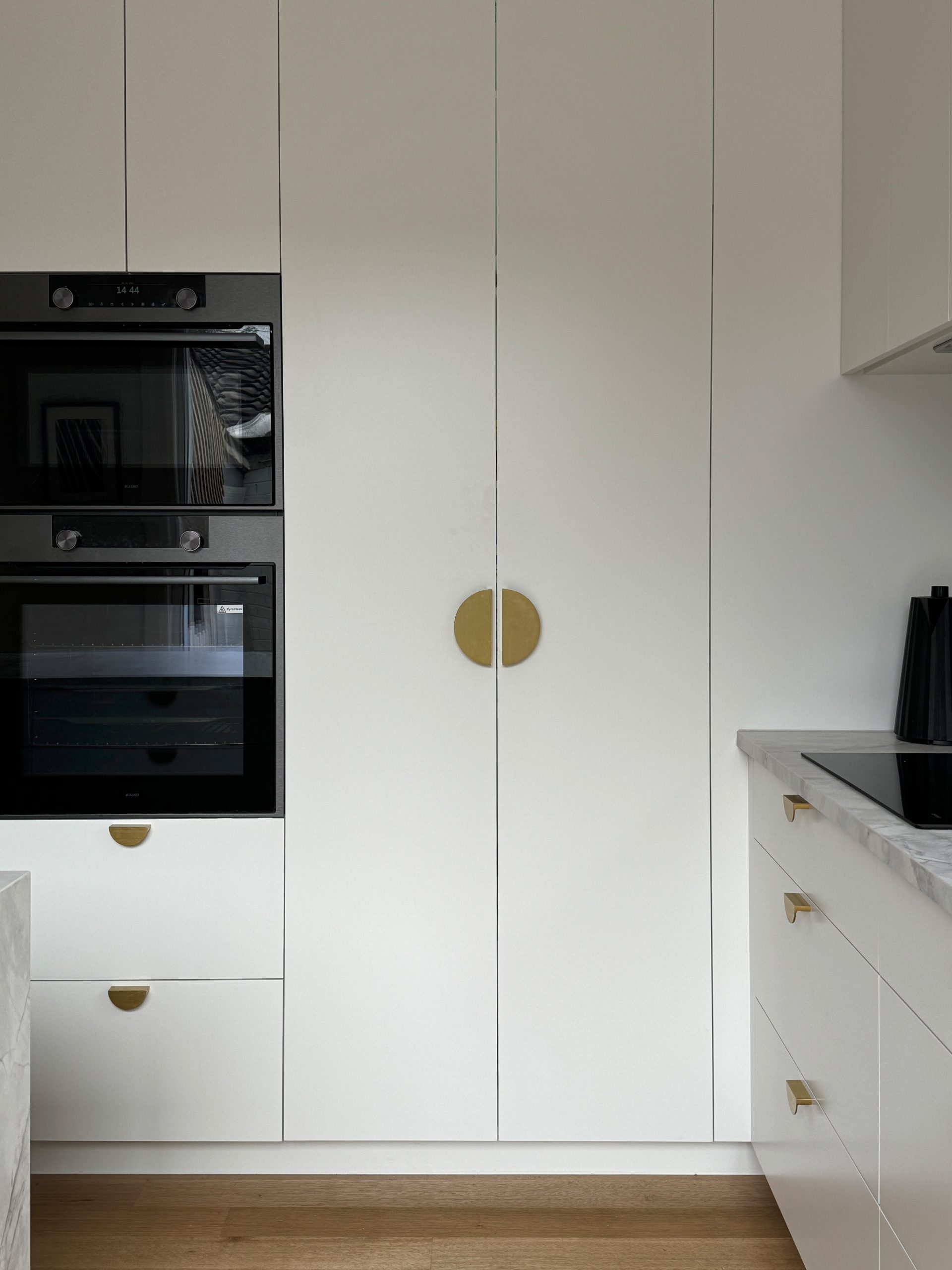 Minimalist kitchen with white cabinets, black oven, and stovetop. Wooden floor and brass handles on doors and drawers. Minimalist kitchen with white cabinets, black oven, and stovetop. Wooden floor and brass handles on doors and drawers.