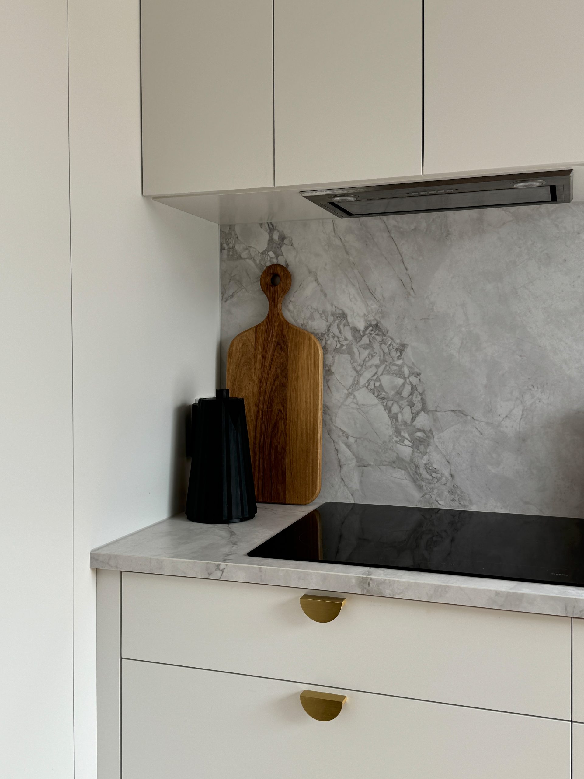 Minimalist kitchen with marble backsplash, wooden cutting board, black containers, and stovetop on white counters. Minimalist kitchen with marble backsplash, wooden cutting board, black containers, and stovetop on white counters.