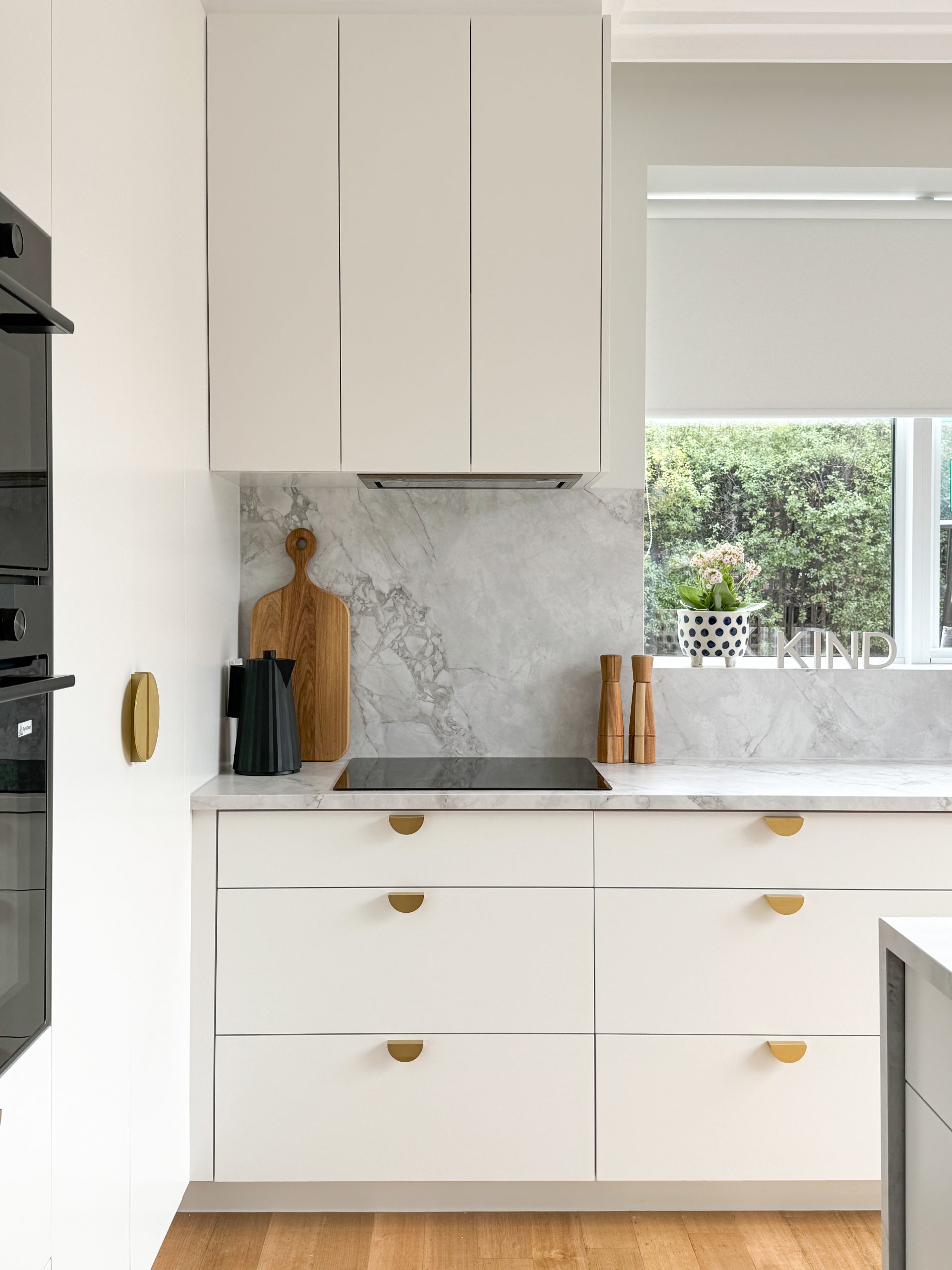 Modern kitchen with white cabinets, gold handles, marble backsplash, black stove, wooden accents, and window view.