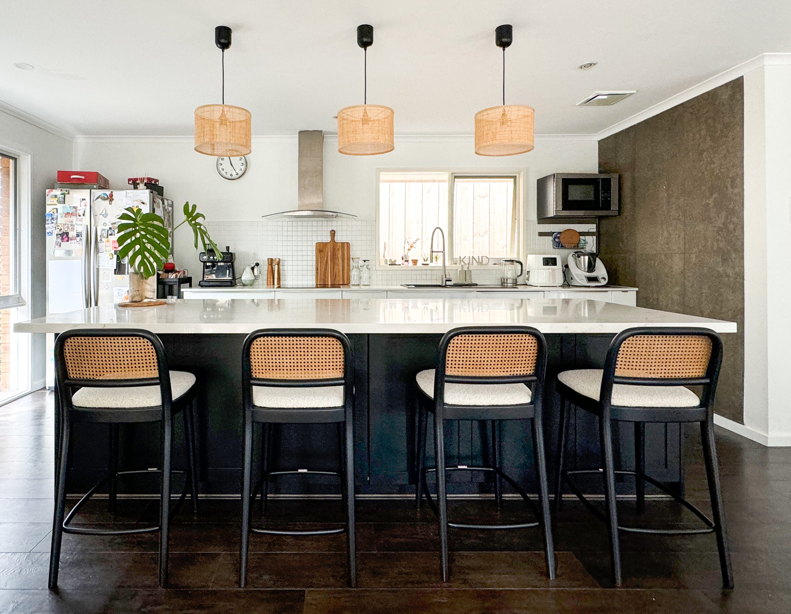 Modern kitchen with a large island, wicker bar stools, hanging lamps, and various appliances and decor on countertops.