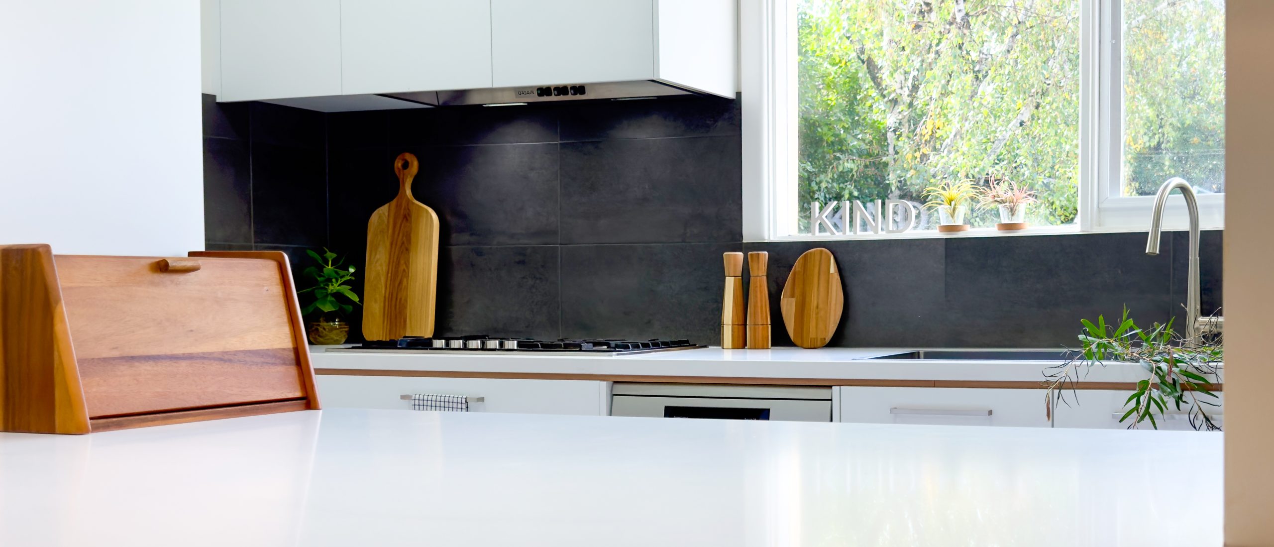 Modern kitchen with dark backsplash, wooden cutting boards, and plants on white countertops near a window. Modern kitchen with dark backsplash, wooden cutting boards, and plants on white countertops near a window.