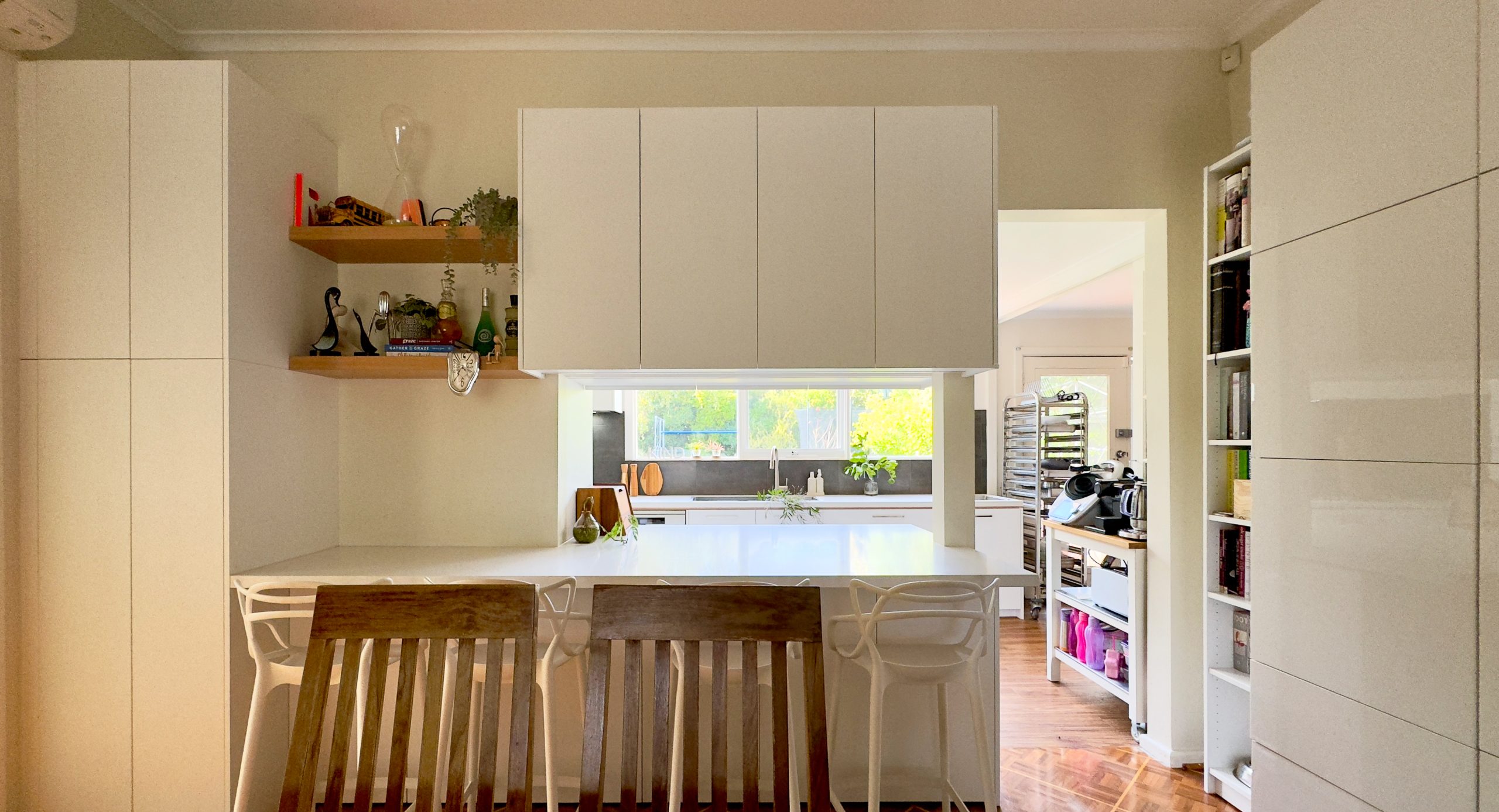 Modern kitchen with white cabinets, wooden chairs, open shelves, and a window with greenery visible.