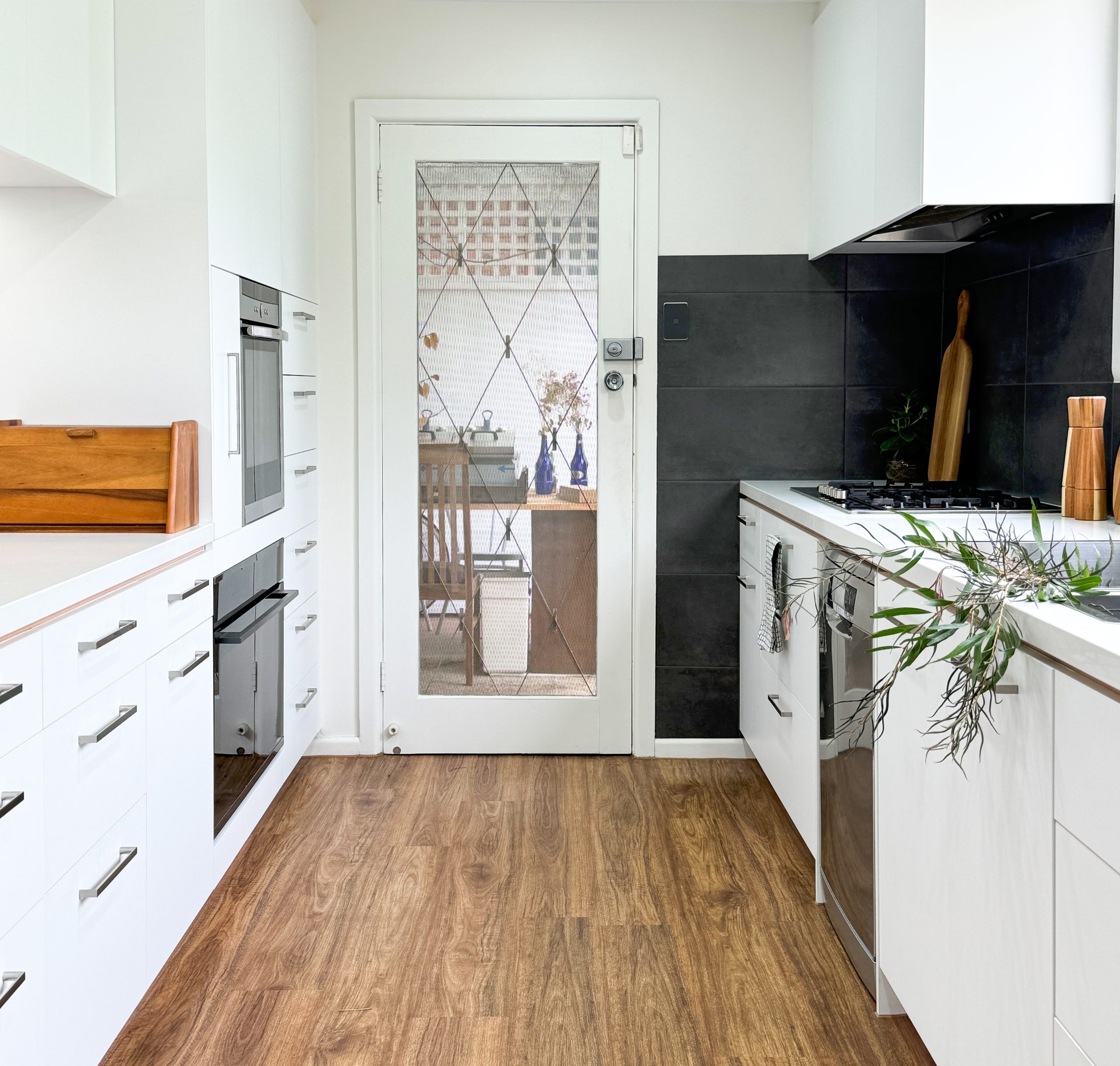 Modern kitchen with white cabinets, wood floor, and glass door showing a dining area outside. Plants and decor on counters. Modern kitchen with white cabinets, wood floor, and glass door showing a dining area outside. Plants and decor on counters.