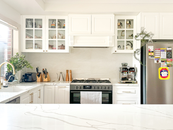 Bright kitchen with white cabinets, a stainless steel fridge, gas stove, and marble countertops. Plants add greenery.