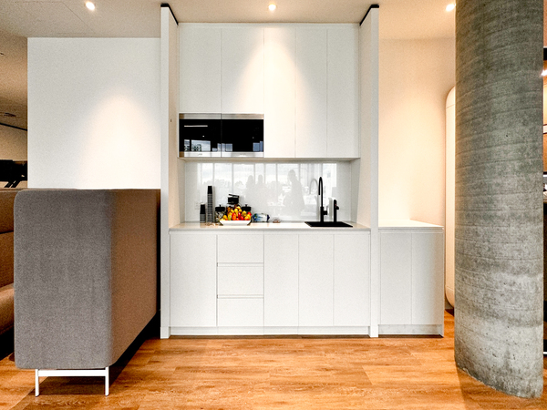 Modern kitchen area with white cabinets, black sink, and a small fruit basket. Light wooden floor and round pillar.