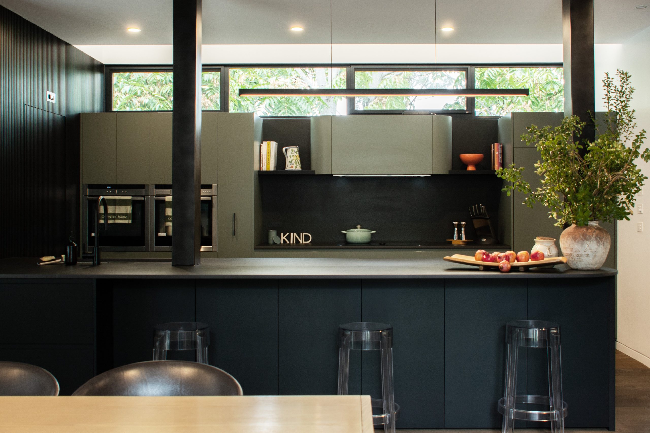 Ben+Mel-Hawthorn-01 A modern, two-tone kitchen featuring a large, dark island with clear acrylic bar stools, set against a back wall with olive-green upper cabinets and black lower cabinetry.